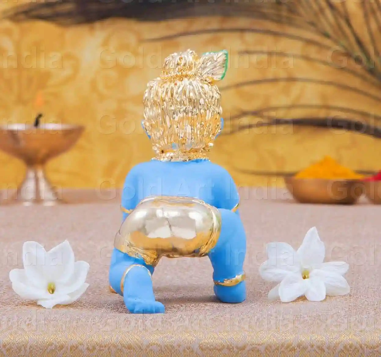 blue-colored Baby Krishna idol in a crawling pose, adorned with gold jewelry, a peacock feather on his head, and holding a golden laddu in his hand
