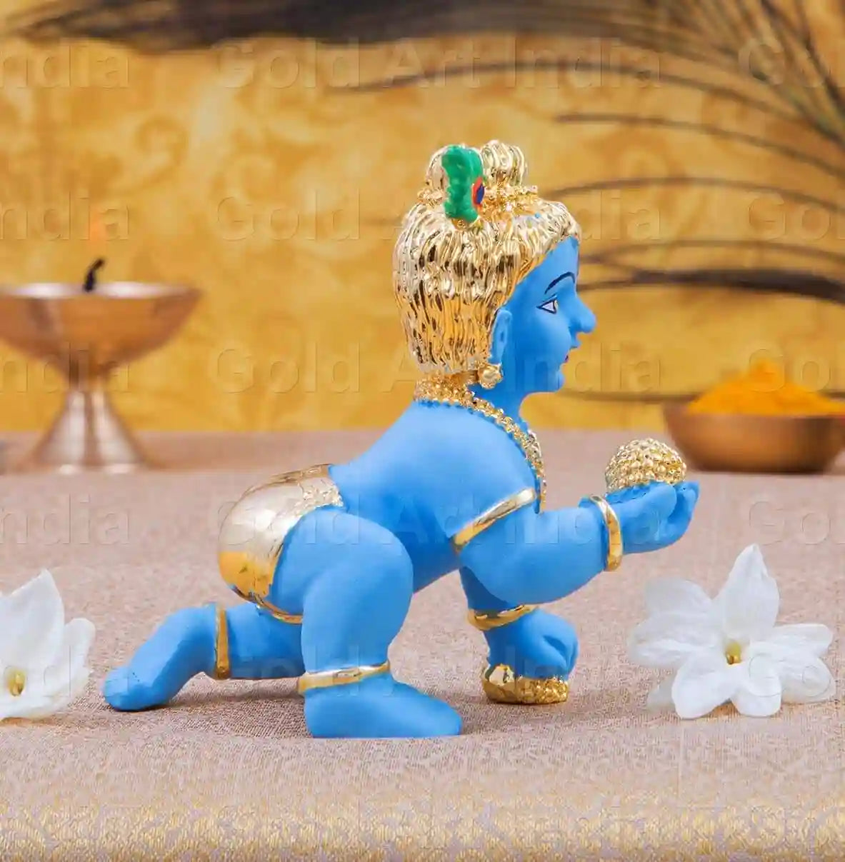 blue-colored Baby Krishna idol in a crawling pose, adorned with gold jewelry, a peacock feather on his head, and holding a golden laddu in his hand