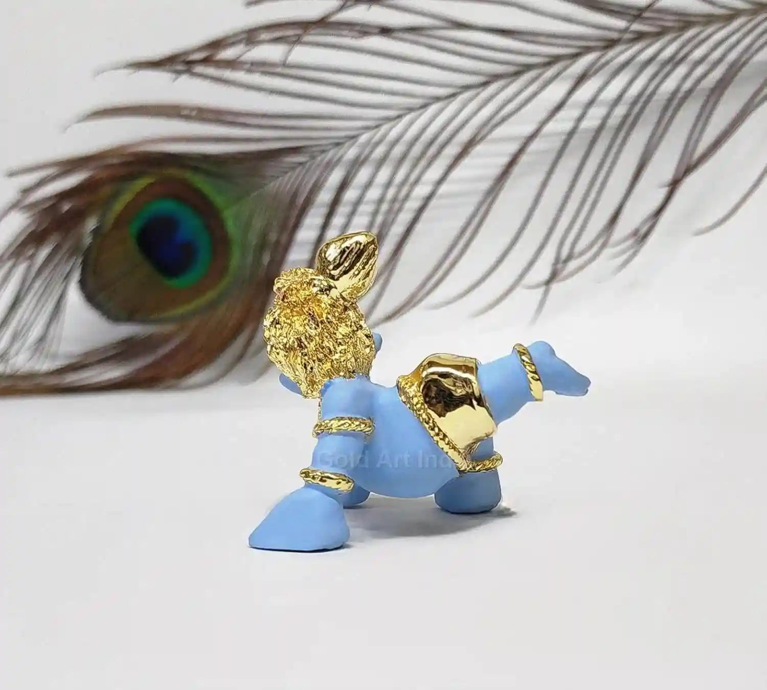 A side view of the baby Krishna idol, with focus on the back details and peacock feather, against a blurred background featuring a peacock feather.
