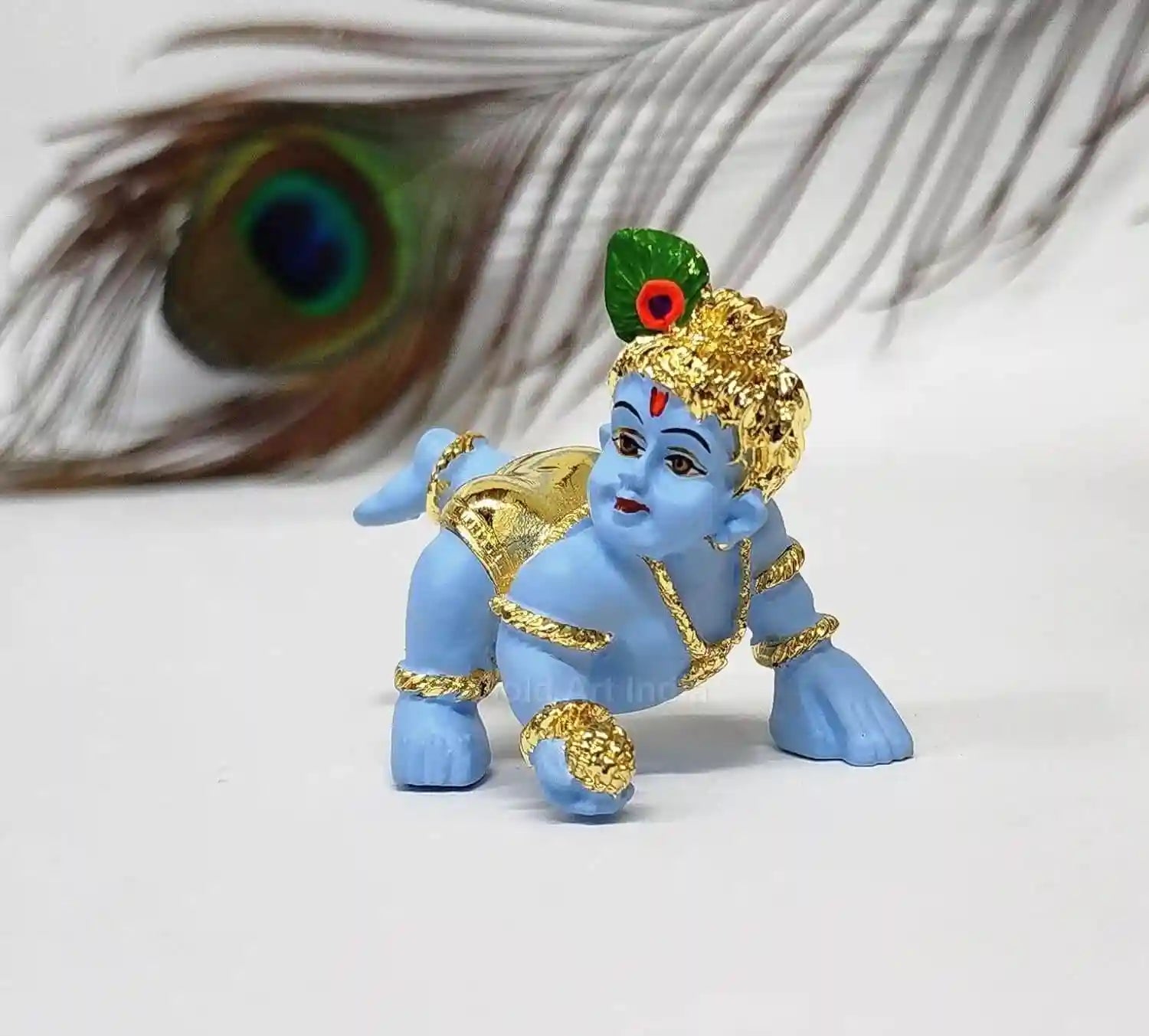 A small blue idol of baby Krishna crawling, adorned with Gold Plated and a peacock feather on his head. The idol is placed in front of a large peacock feather.