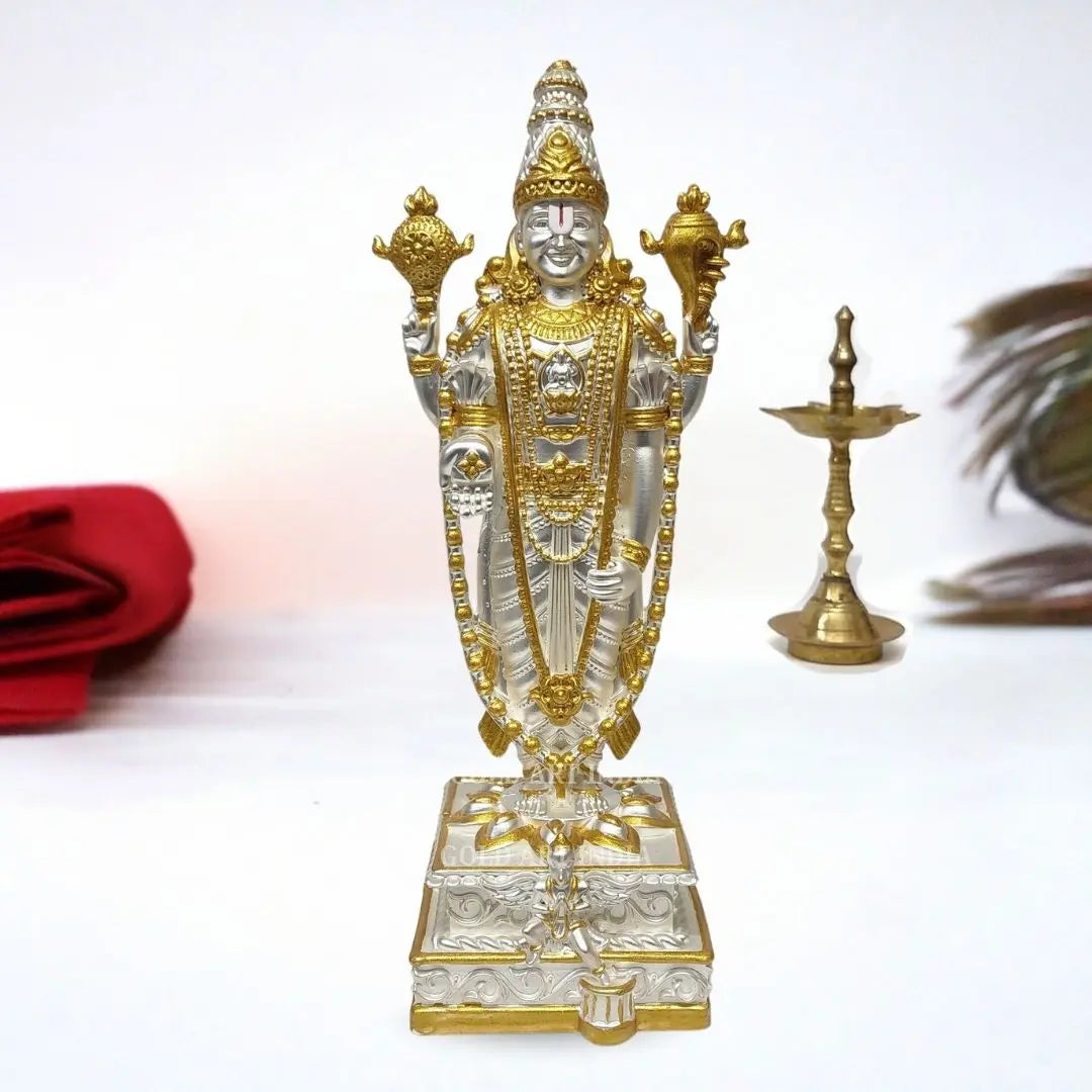 Silver-and-gold Plated Lord Venkateswara statue on a pedestal with a brass lamp in the background.
