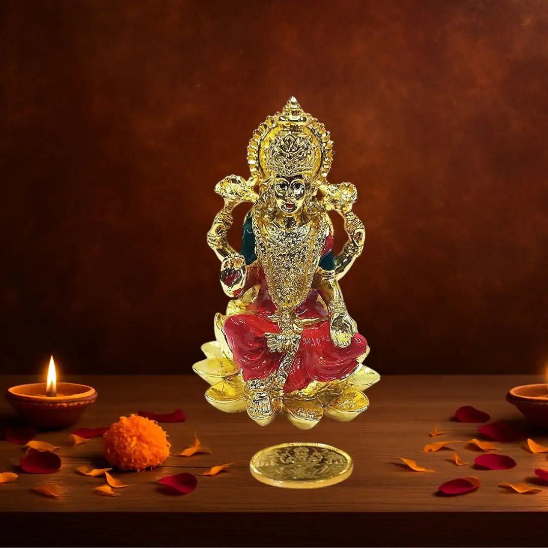 Gold plated Kamal Lakshmi idol with orange enamel detailing, seated on lotus base, accompanied by gold-plated coin, decorated with diya and flowers.