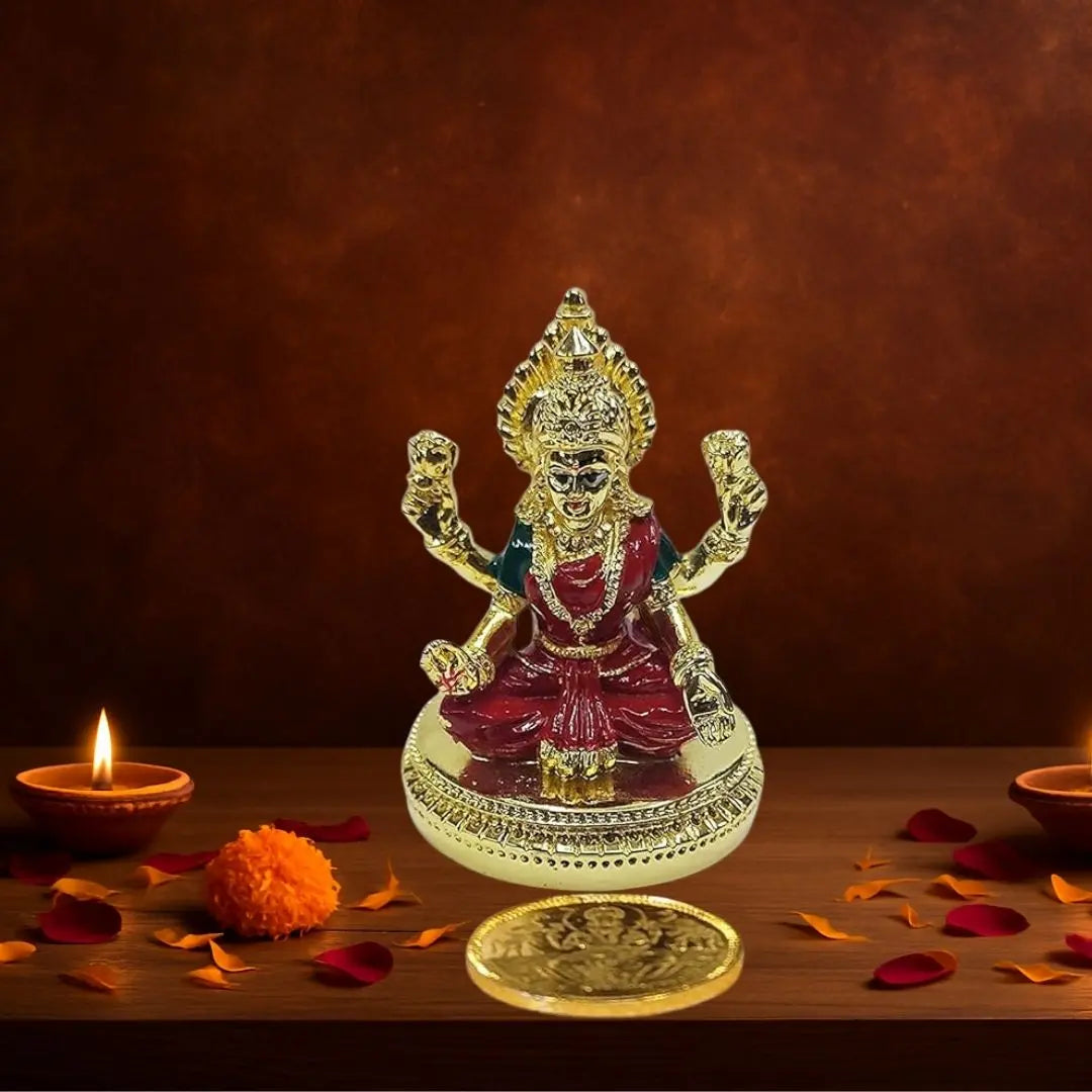 Gold plated Lakshmi idol with red enamel detailing, seated on round base, accompanied by gold-plated coin, decorated with diya and flowers.