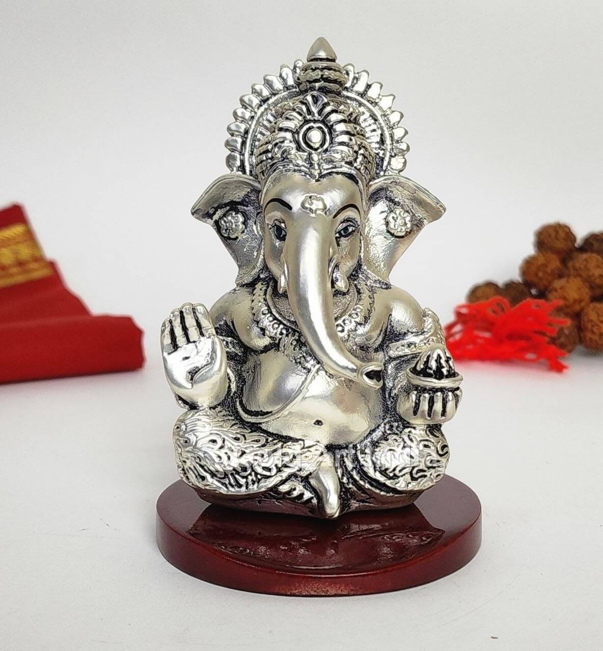 A 3.5-inch Silver Plated Antique Finish Mdf base Ganesha statue with a crown, jewelry, and intricate carvings. One hand gives blessings, the other holds a modak. Decorated with Rudraksh and red cloth in the background.