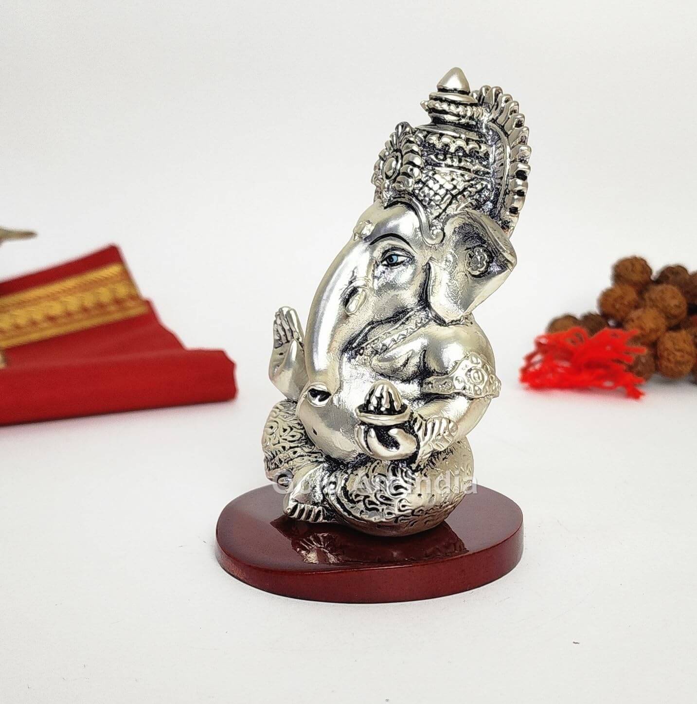 A 3.5-inch Silver Plated Antique Finish Mdf base Ganesha statue with a crown, jewelry, and intricate carvings. One hand gives blessings, the other holds a modak. Decorated with Rudraksh and red cloth in the background. side view