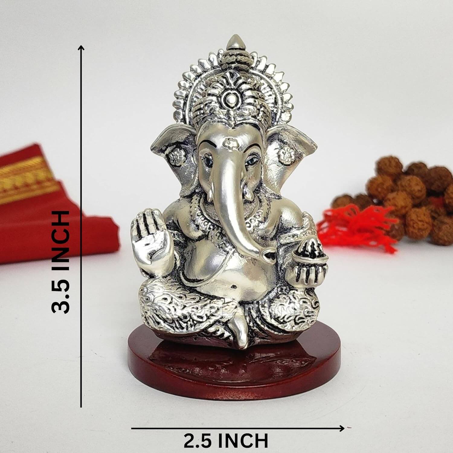 A 3.5-inch Silver Plated Antique Finish Mdf base Ganesha statue with a crown, jewelry, and intricate carvings. One hand gives blessings, the other holds a modak. Decorated with Rudraksh and red cloth in the background.