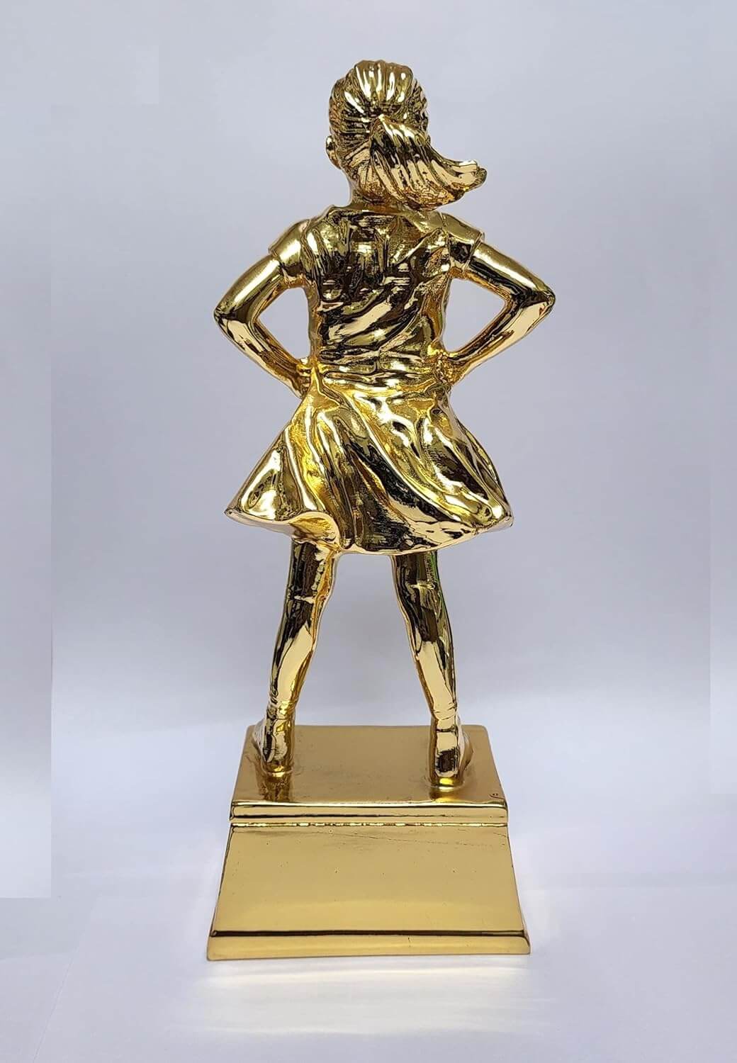 A Gold -plated trophy featuring a young girl standing confidently with her hands on her hips. This is a rear view of the trophy, showing the flowing details of her dress and her neatly tied ponytail. The figure stands on a silver rectangular base. The background is a plain white surface.