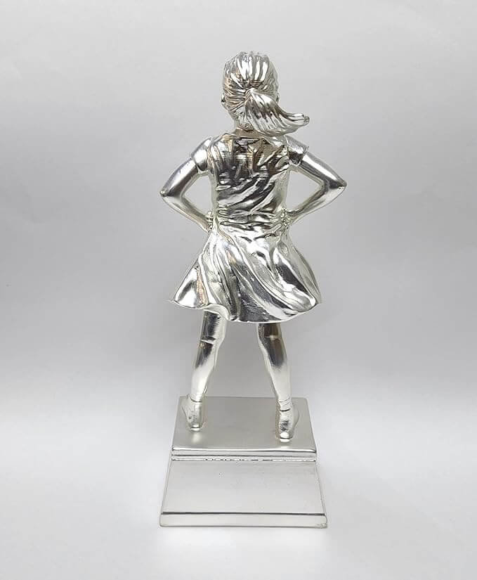 A silver-plated trophy featuring a young girl standing confidently with her hands on her hips. This is a rear view of the trophy, showing the flowing details of her dress and her neatly tied ponytail. The figure stands on a silver rectangular base. The background is a plain white surface.