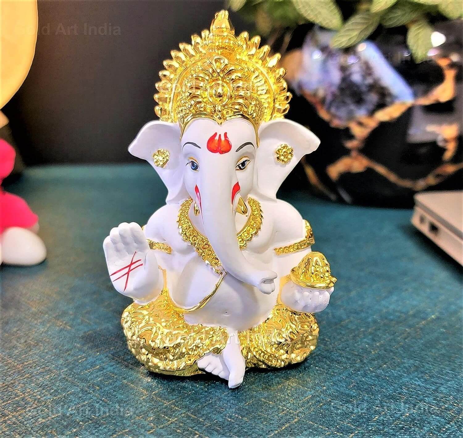 A beautifully crafted white and gold Lord Ganesha idol placed on a table, surrounded by decorative elements like a plant, a miniature bullock cart, and a laptop.
