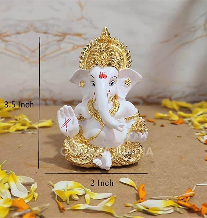 A white and gold Lord Ganesha idol measuring 3.5 inches in height and 2 inches in width, surrounded by yellow and orange flower petals.