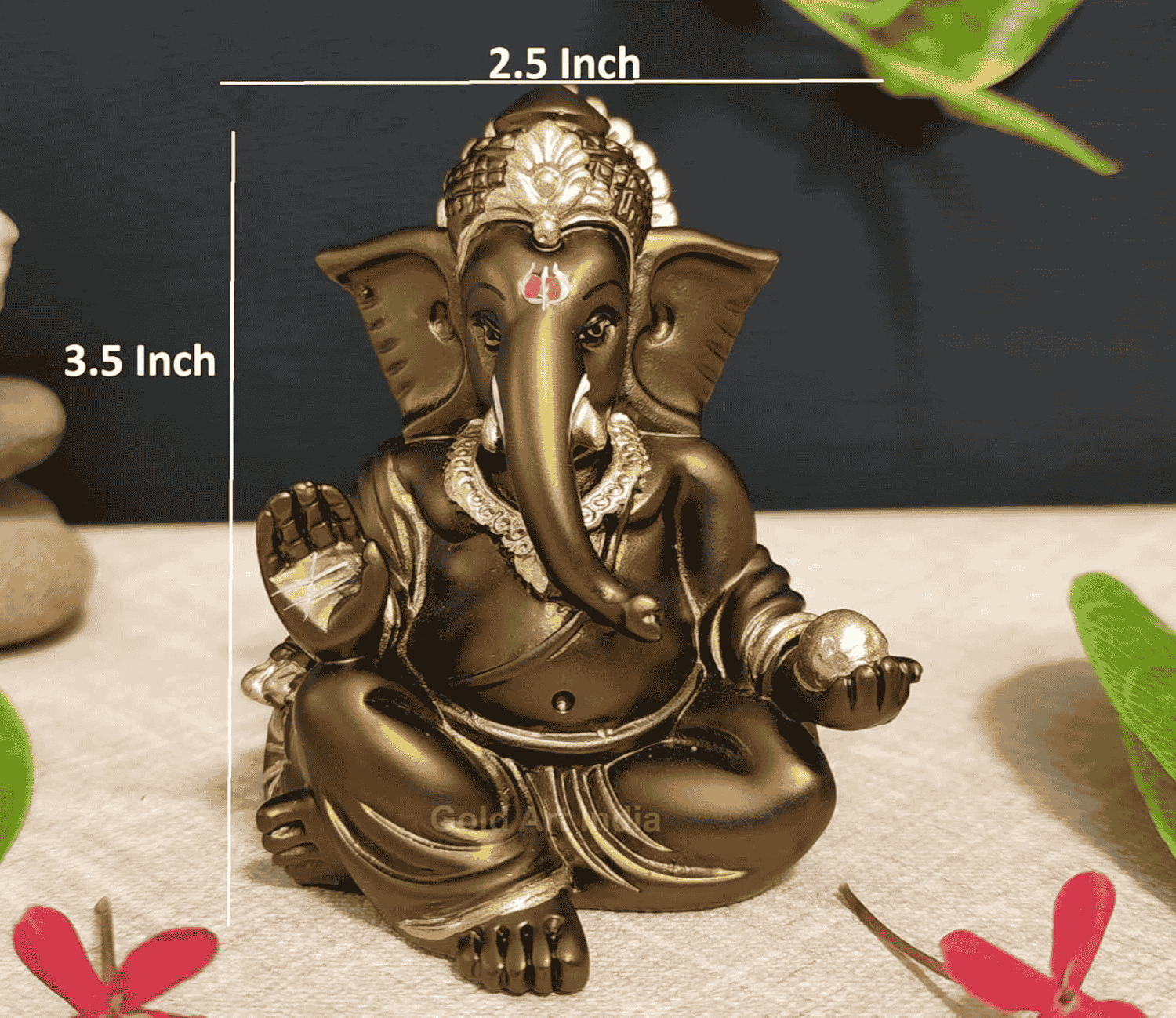 A 3.5-inch bronze-colored Ganesha statue with a crown, jewelry, and intricate carvings. One hand gives blessings, the other holds a golden Ladoo. Decorated with green leaves and red flowers in the background.