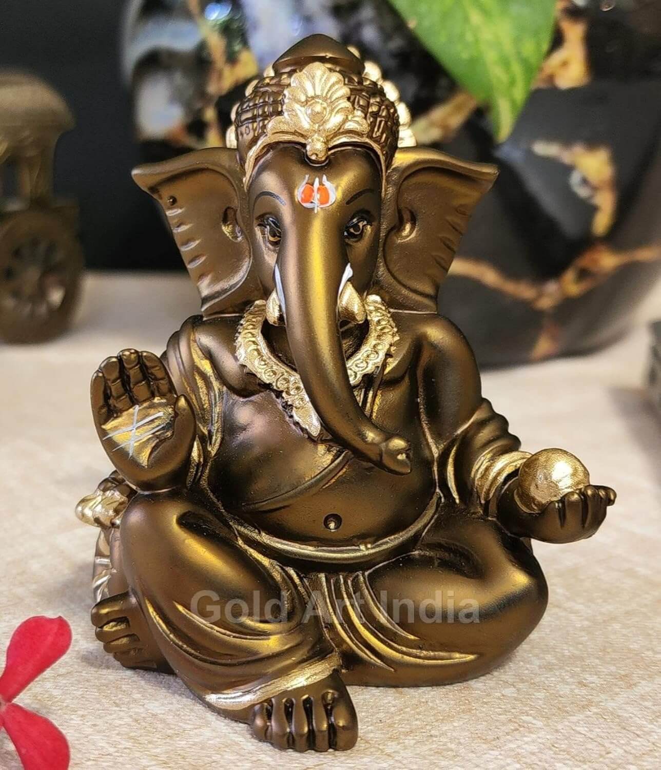 A 3.5-inch bronze-colored Ganesha statue with a crown, jewelry, and intricate carvings. One hand gives blessings, the other holds a golden Ladoo. Decorated with green leaves and red flowers in the background.