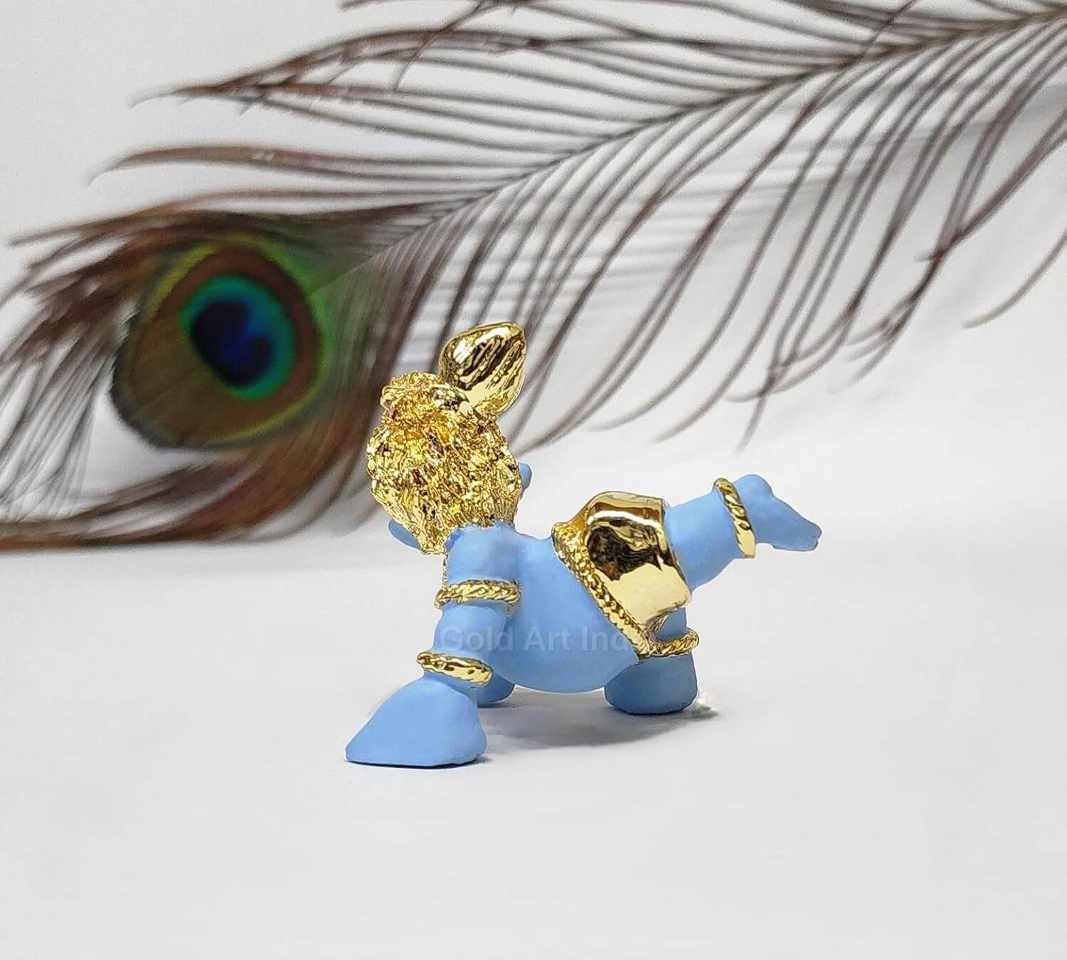 A side view of the baby Krishna idol, with focus on the back details and peacock feather, against a blurred background featuring a peacock feather.