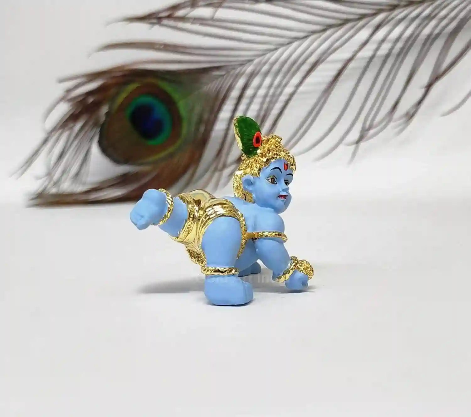A side view of the baby Krishna idol, with focus on the back details and peacock feather, against a blurred background featuring a peacock feather.