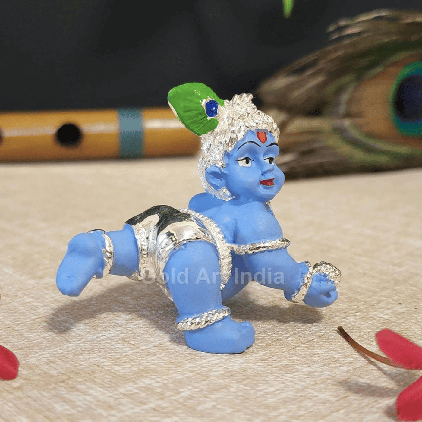 A close-up of the baby Krishna idol, showing fine details of its design. A flute and a peacock feather are placed in the background.