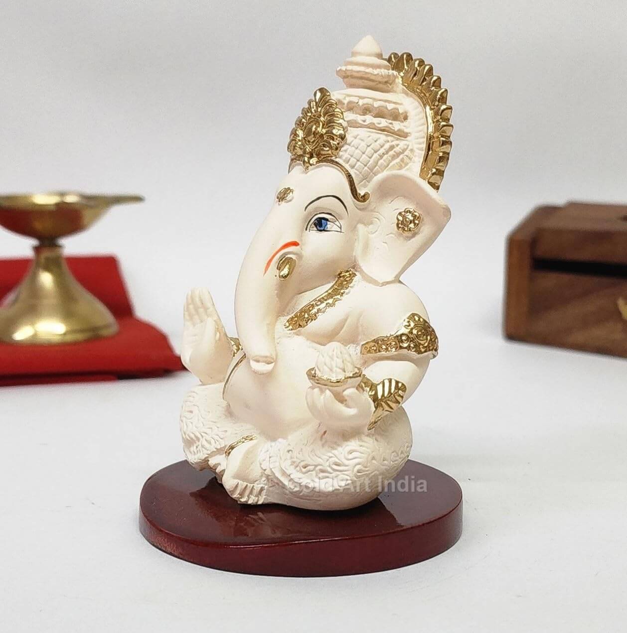 A 3.5-inch Peach Ivory Mdf base  Ganesha statue with a crown, jewelry, and intricate carvings. One hand gives blessings, the other holds a modak. Decorated with box and red cloth in the background.
